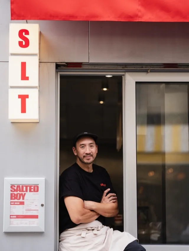 Assis à l'entrée de la boutique, tablier noué, sous l'enseigne SLT et le store rouge.