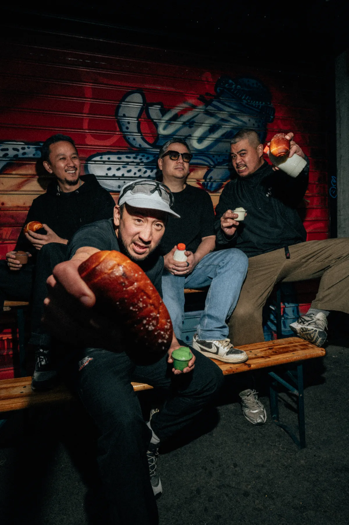 Le crew Salted Boy sur des bancs devant un rideau de fer graffé, salted bread tendu vers l'objectif.